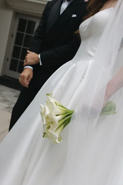 Bridal portrait of a bride holding a bouquet of ivory calla lilies by a glass door on a stone floor.