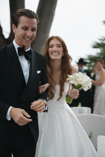 Ceremony exit during a newlywed aisle walk, bride and groom smiling as she carries a white calla lily bouquet, joyful and candid.
