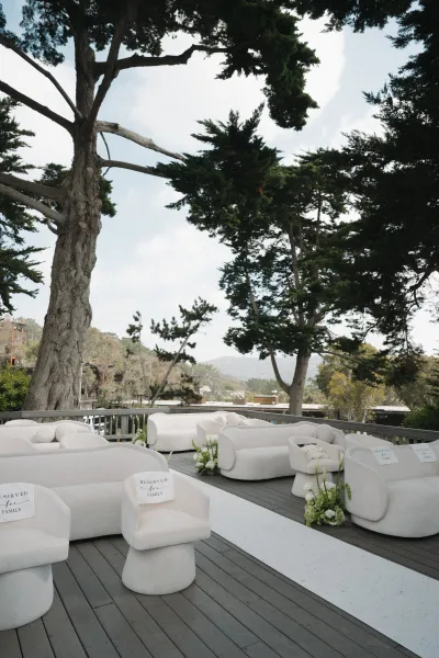 Ceremony seating styled as lounge ceremony seating with white sofas on a wood deck, bordered by evergreen trees and distant hills.