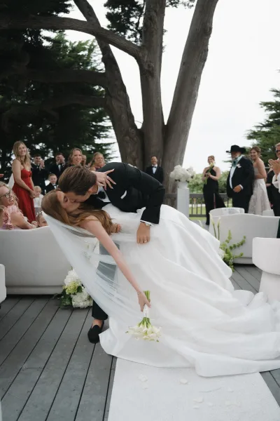 Wedding kiss portrait with a dip kiss as the groom lowers the bride, her veil flowing, on a wooden deck beneath a large garden tree.