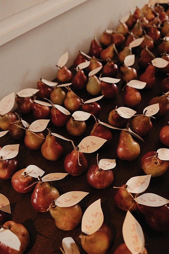 Wedding place cards displayed on pears with paper tags and twine, arranged on a rustic wood table against a white wall