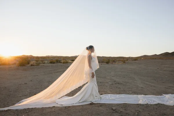 Bridal portrait of a bride with long veil, standing in golden sunset light across a quiet desert plain with distant hills.