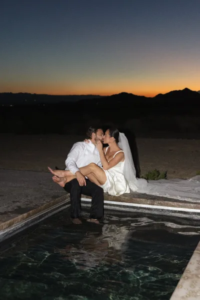 Wedding kiss portrait of a poolside newlywed couple, her veil cascading, with desert mountains and a sunset sky in the background.