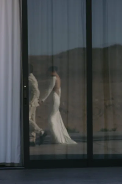 Couple portrait of a bride and groom walking hand in hand, her bridal gown catching the light by glass doors with desert hills beyond on a patio.