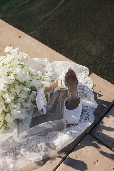Wedding shoes—white bridal heels with clean lines—arranged on stone paving by shimmering pool water, softened by a trailing bridal veil.