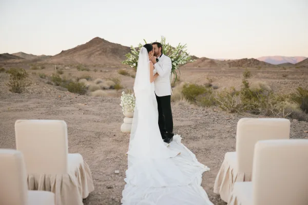 Wedding first kiss as a couple at the altar, her cathedral bridal veil trailing while desert mountains and shrubs stretch beneath a wide sky.