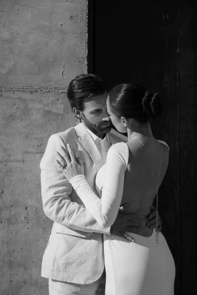 Black and white couple portrait of a bride and groom embrace, near a kiss, her open-back gown showing a back necklace against concrete and wood.