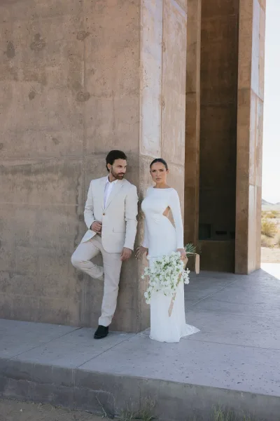 Couple portrait with bride holding bouquet as the groom leans in a tan suit, set against a concrete wall, desert landscape, and wide blue sky.