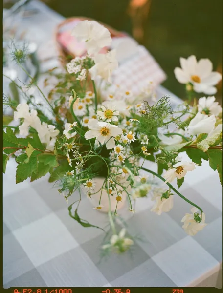 Wedding tablescape with a wildflower wedding centerpiece of white blooms and greenery on a gingham tablecloth, set on a sunlit lawn