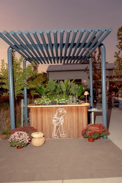 Wedding bar setup with an outdoor wedding bar under a blue pergola, featuring glassware, greenery, white flowers, and pumpkins on a patio at dusk