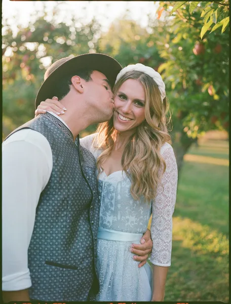 Wedding couple portrait with groom kissing bride’s cheek as she smiles at camera, headband accent, in a sunset-lit orchard lawn