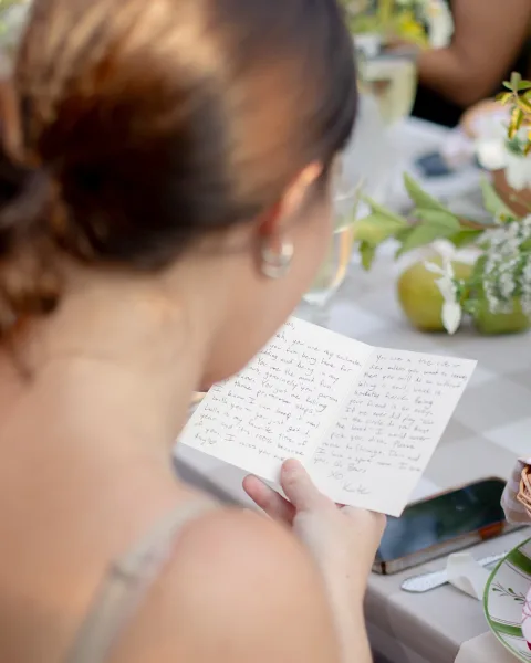 Wedding letter reading as the bride reads a handwritten note at the reception table, pearl earrings and champagne flute beside florals and fruit