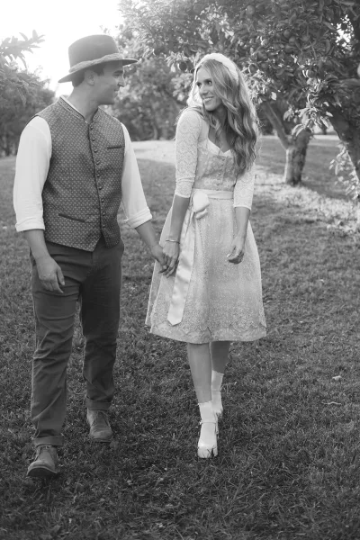 Couple portrait of bride and groom walking hand in hand, her lace dress with sash and his hat, among orchard trees on grass lawn