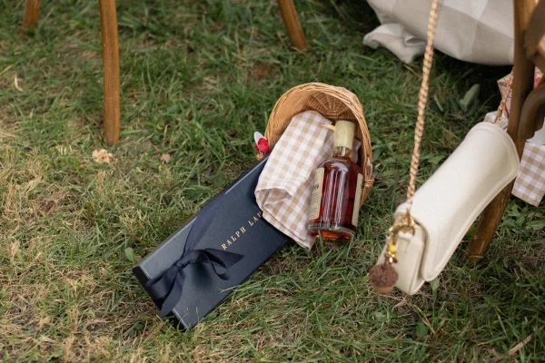 Wedding welcome bag in a wicker basket with a liquor bottle, gift box, and gingham cloth on a grass lawn near chair legs
