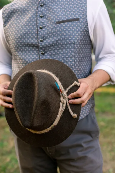 Groom accessories featuring a brown felt hat with rope band and feather pin, paired with a patterned gray vest over white shirt outdoors