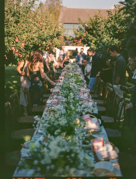 Outdoor reception table set as a long banquet table wedding with floral garland, white blooms, candles, and wooden chairs under apple trees