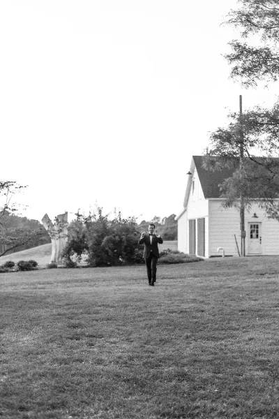 Groom portrait of a man in a tuxedo with a bow tie, standing on a grass lawn with trees and a farmhouse building behind him