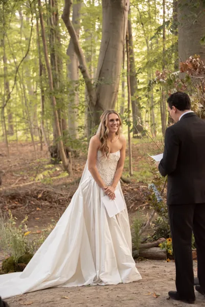 Wedding vows as groom reads from vow papers while bride in strapless lace dress and veil listens on a leafy forest path