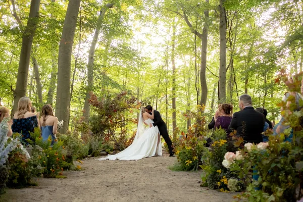 Ceremony kiss moment as bride in veil and strapless dress kisses groom in black suit on a forest aisle with grounded flowers