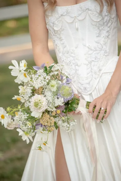 Bridal bouquet of white cosmos and wildflower wedding bouquet with baby’s breath, held by bride in beaded dress on a garden walkway