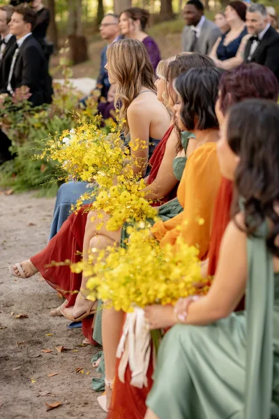 Wedding guests seated at an outdoor ceremony guests in a wooded setting, bridesmaids holding bright yellow bouquets with ribbons along the dirt aisle
