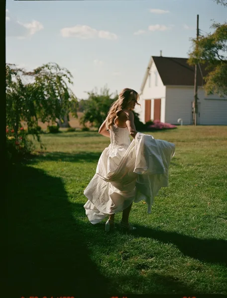 Bridal portrait of a bride holding dress, showing a corset back gown and layered skirt on a sunny lawn near a rustic farmhouse