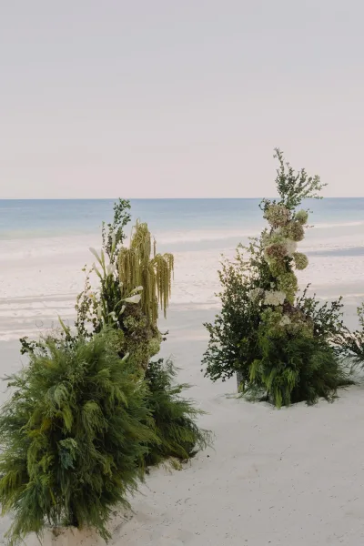 Beach ceremony decor with tropical wedding ceremony flowers on two floral pillars of hydrangeas, calla lilies, greenery, and hanging amaranthus by the ocean horizon