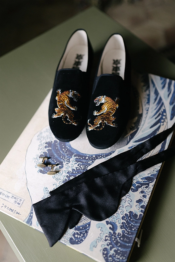Groom accessories arranged in a flat lay with black embroidered loafers, cufflinks, black bow tie, and a printed book on a green chair indoors