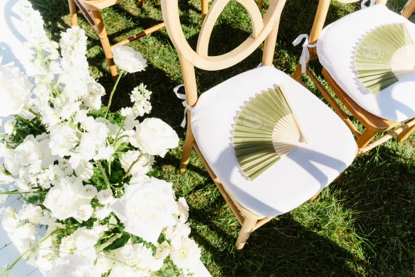 Ceremony aisle decor with wooden chairs and white cushions, paper hand fans, and white rose florals lining an aisle runner on a sunlit lawn