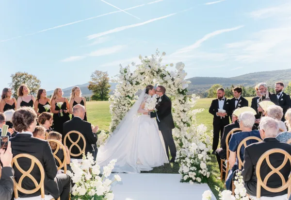 Ceremony kiss as bride and groom embrace beneath a round white floral arch, long veil flowing on a mountain lawn aisle with guests seated