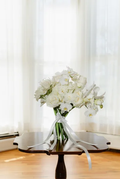 Bridal bouquet of white roses and orchids with cascading ribbon tails and greenery on a wood table in soft window light