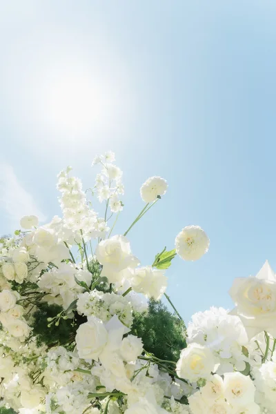 Wedding florals of white wedding flowers with roses, hydrangea, carnations, baby’s breath and greenery against a blue sky with sun flare