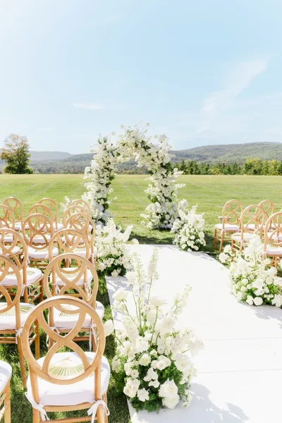 Ceremony setup with an outdoor wedding ceremony floral wedding arch, white aisle runner and chairs on a mountain-view lawn under blue sky