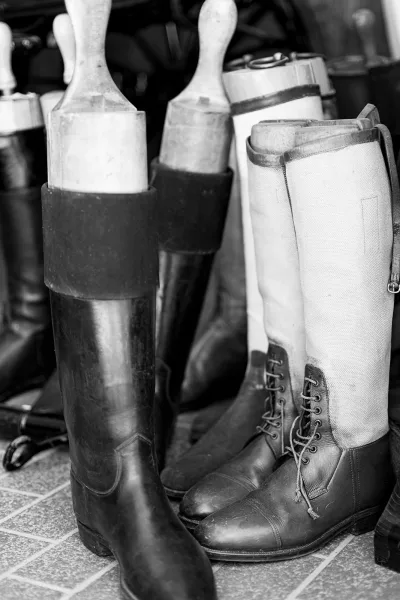 Riding boots and tall leather boots lined up with boot trees on a tile floor in a dark interior, showing polished leather and laces