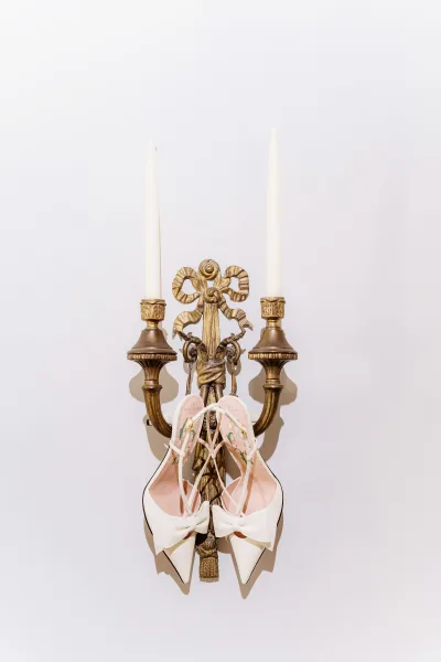 Bridal shoes with white bow heels hang from a vintage brass wall sconce, framed by taper candles against a clean white wall