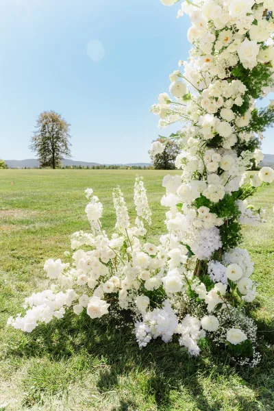 Wedding ceremony arch with white floral ceremony arch of roses and baby’s breath, asymmetrical greenery framing a grassy field under blue sky