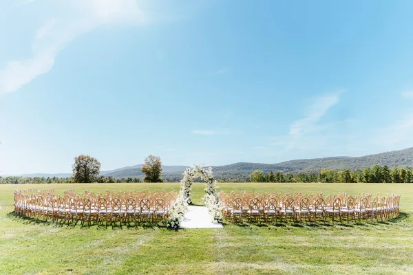 Ceremony setup with an outdoor wedding ceremony floral arch, white aisle runner, and wooden chairs on a mountain-view lawn under blue sky