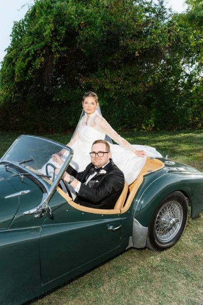 Couple portrait of bride and groom in a vintage wedding car, her lace sleeves and veil visible as he drives past leafy trees