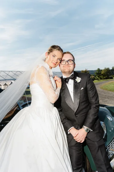 Couple portrait of bride in lace sleeves leaning on groom in black tuxedo by a classic car on a sunny gravel driveway near an event tent