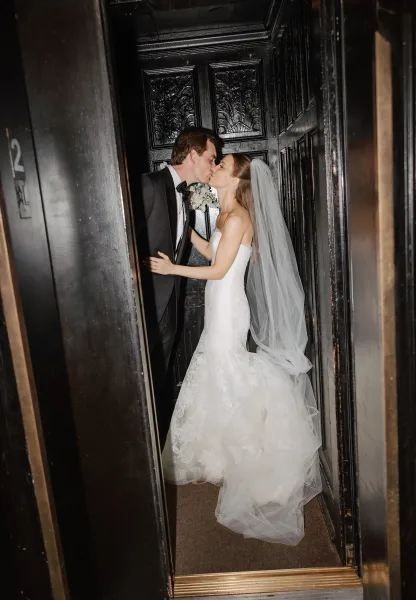 Wedding kiss portrait of bride and groom kissing in a narrow hallway by a dark wood door, her cathedral veil trailing and bouquet raised