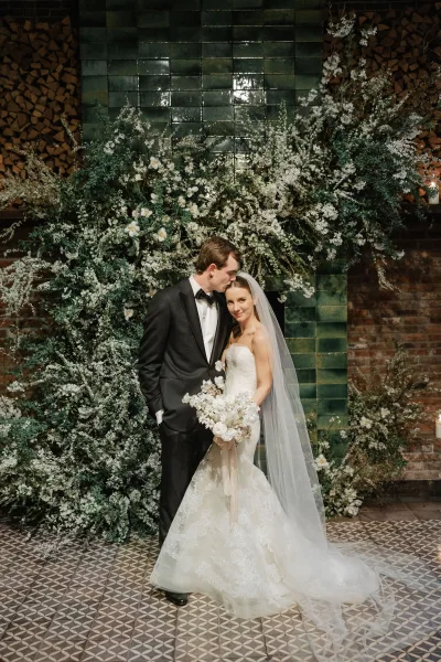 Couple portrait with groom kissing bride’s forehead as she holds a white orchid bouquet, framed by candles and floral wall on green tile backdrop