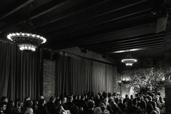 Wedding ceremony as bride in veil and groom in suit stand at floral altar with candles beneath chandelier in brick-walled loft with guests seated