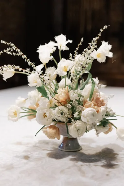 Wedding centerpiece with white and peach tulips in a compote vase, accented by ranunculus and baby's breath on linen tablecloth against dark backdrop