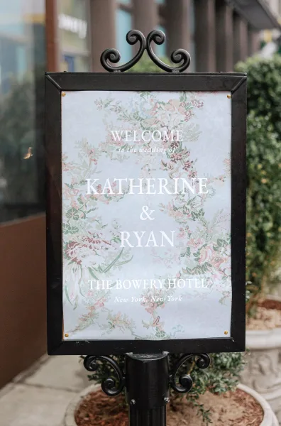 Wedding welcome sign with pastel floral illustration and white lettering in a black frame on a metal stand outside a hotel entrance