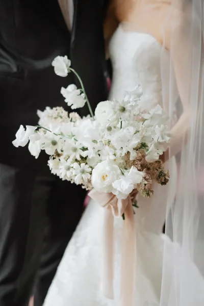 Bridal bouquet of white flowers tied with a blush ribbon, held by a bride in a lace wedding dress beside groom with boutonniere