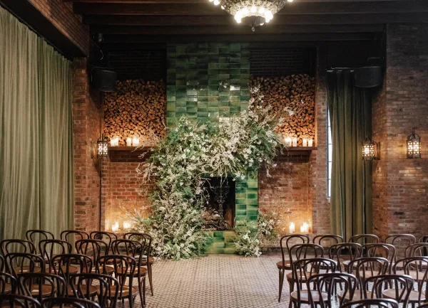 Ceremony backdrop with fireplace wedding altar, asymmetrical white flowers and greenery, candlelit lanterns beneath a chandelier in a brick room