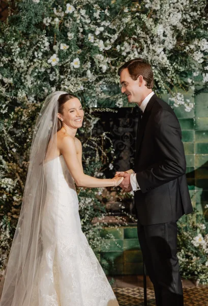 Ceremony moment of bride and groom holding hands, smiling in a black tie indoor wedding ceremony before a floral backdrop and fireplace