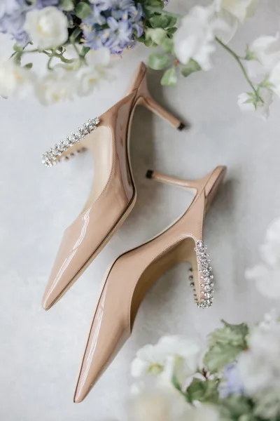 Wedding shoes in nude patent leather with rhinestone ankle straps, styled with white and lavender flowers on a light gray flatlay