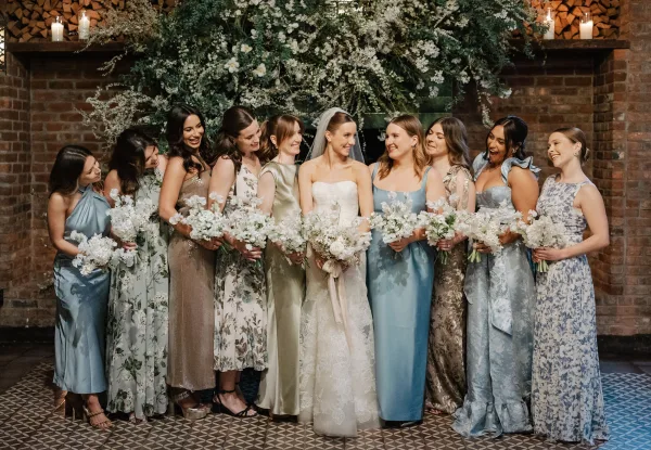 Bridesmaid group photo with bride and bridesmaids in mixed dresses holding white and green bouquets before a candlelit brick fireplace