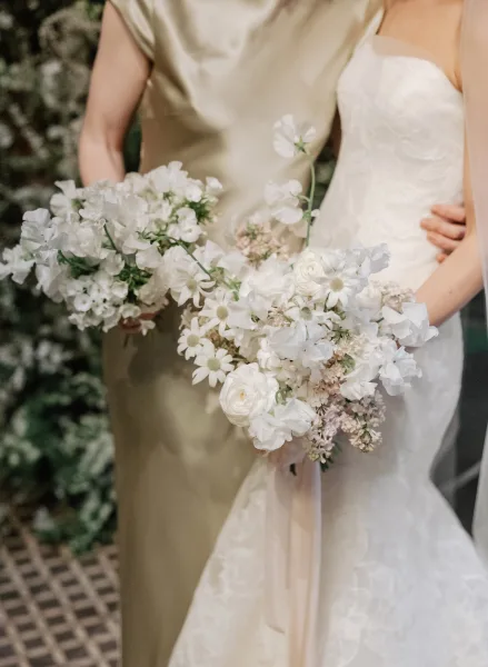Bridesmaid bouquets of white flowers with ribbon streamers held beside bridesmaid dress and bridal gown against a greenery wall
